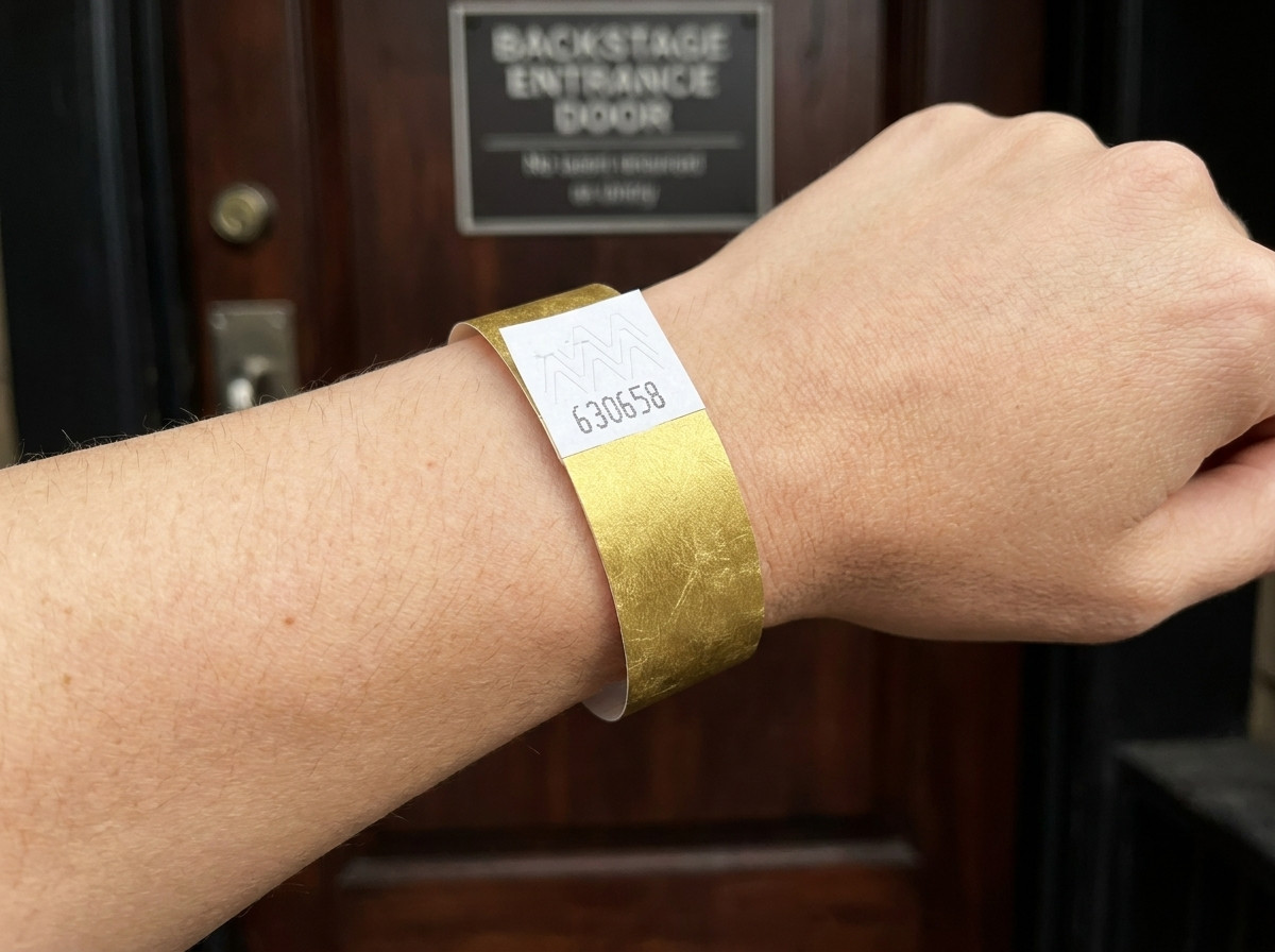 VIP festival ID wristband with gold metallic print and barcode, worn on a wrist in front of a backstage entrance