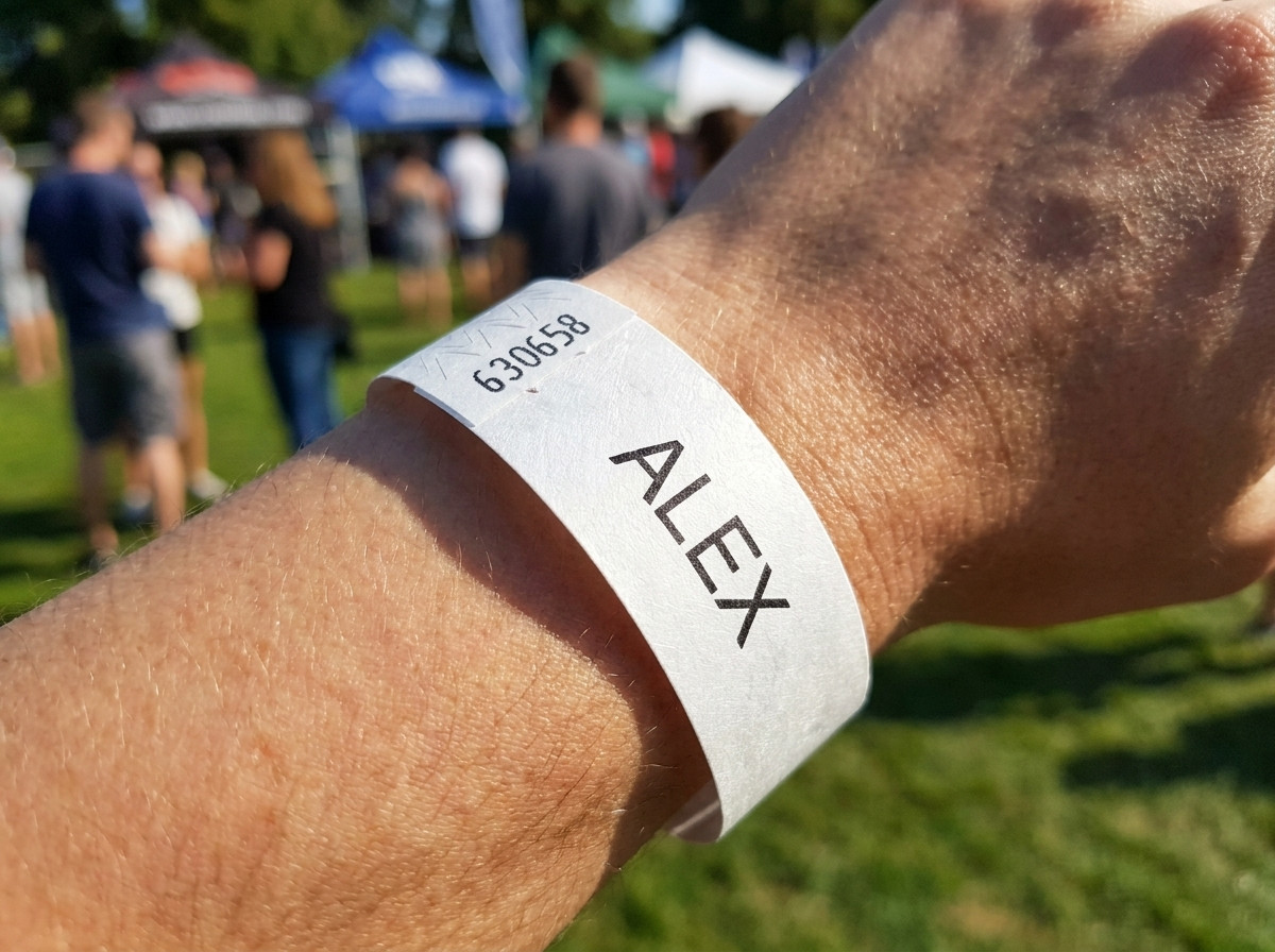 White Tyvek wristband printed with a childs first name and an emergency phone number, worn on a young childs wrist at an outdoor event